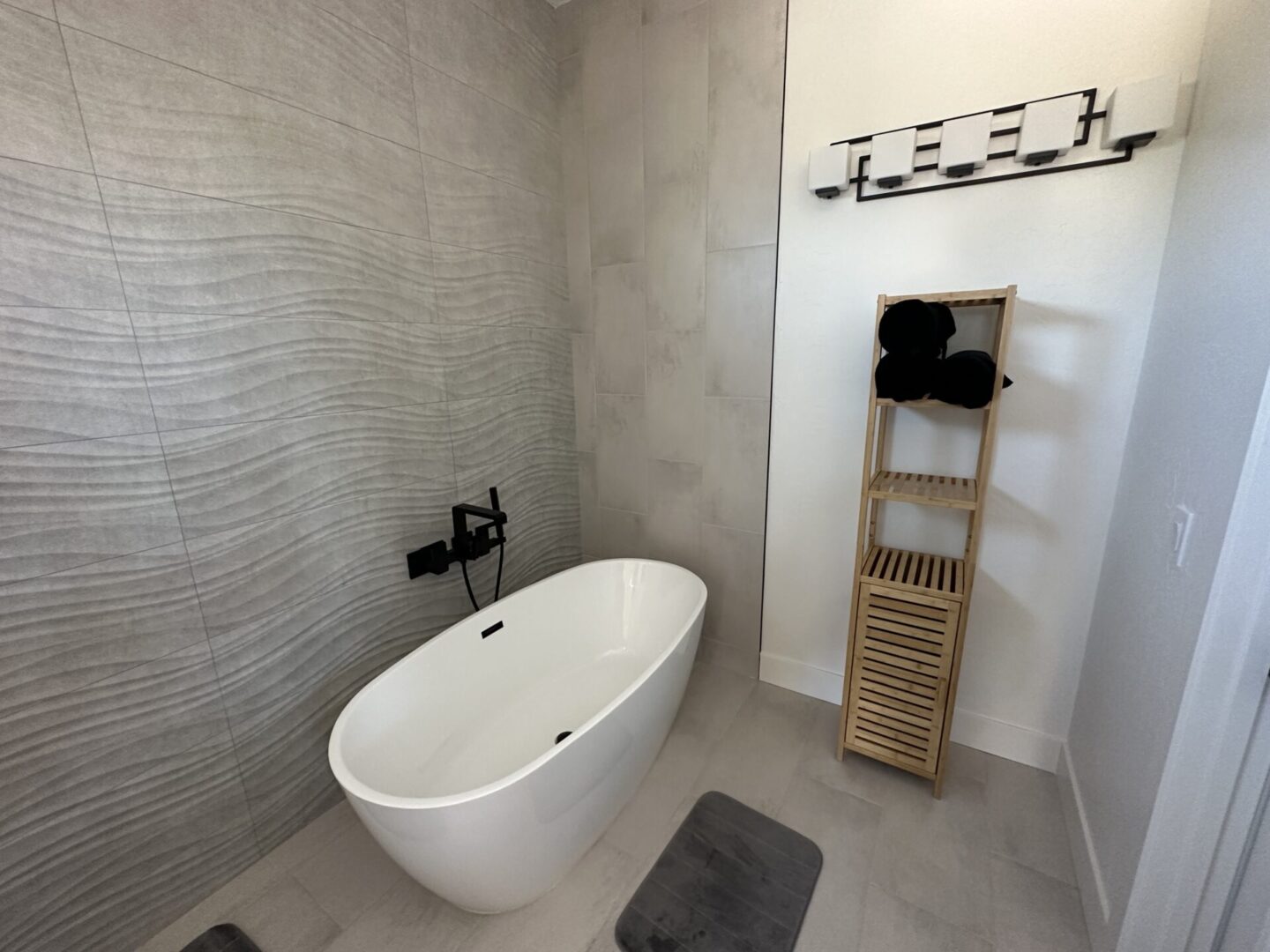 Modern bathroom with freestanding white soaking tub, gray wave-patterned tile surround, wooden towel ladder, chrome fixtures, and light gray flooring