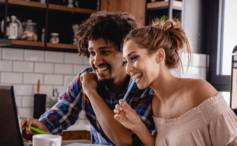 Two people smiling while working together at a desk with a laptop, papers, and colorful markers in a modern kitchen office space