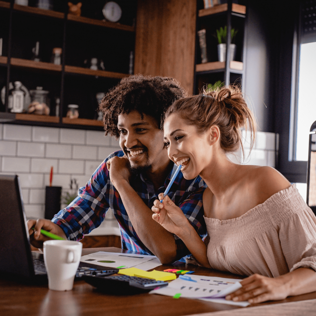 Two people smiling while working together at a desk with a laptop, papers, and colorful markers in a modern kitchen office space
