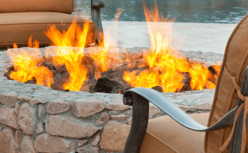 Outdoor fire pit with bright orange flames surrounded by stone wall, tan cushioned chairs facing a beach and water in background