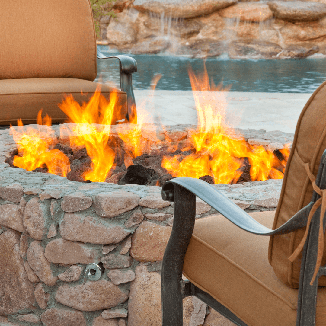 Outdoor fire pit with bright orange flames surrounded by stone wall, tan cushioned chairs facing a beach and water in background