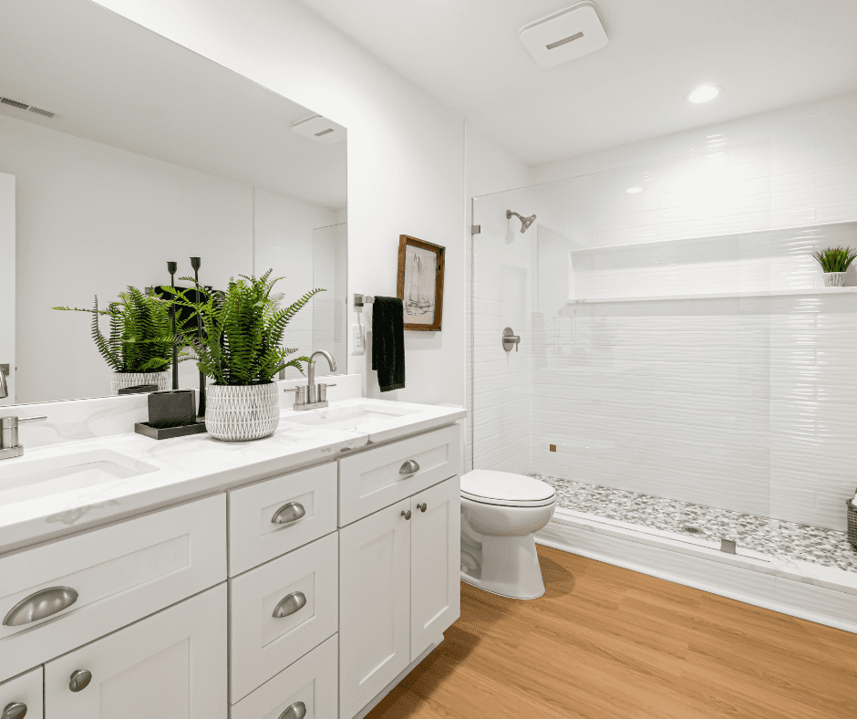 Modern white bathroom with dual vanity, marble countertop, large mirror, walk-in shower with white subway tile, wooden flooring, and potted green plants