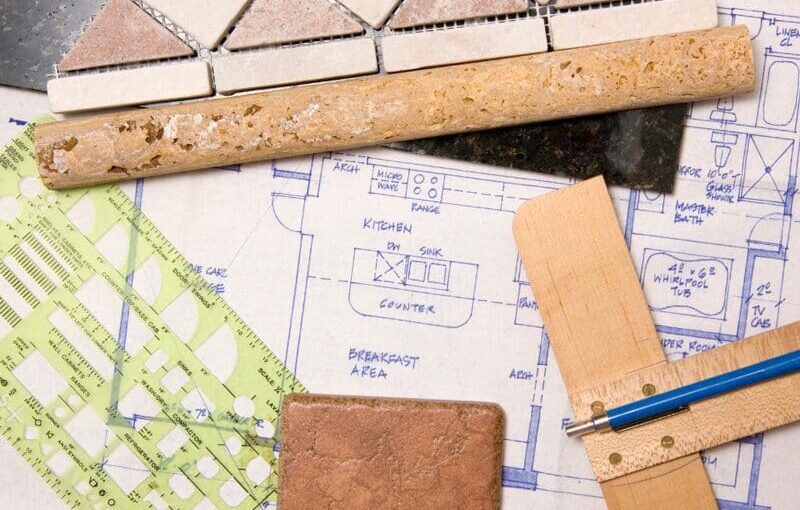 Architectural floor plan with kitchen layout surrounded by design tools including cork ruler, wooden scale, paint samples, green grid ruler, and blue pencil