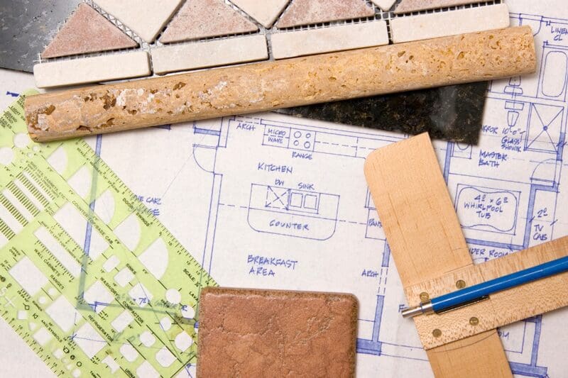 Architectural floor plan with kitchen layout surrounded by design tools including cork ruler, wooden scale, paint samples, green grid ruler, and blue pencil