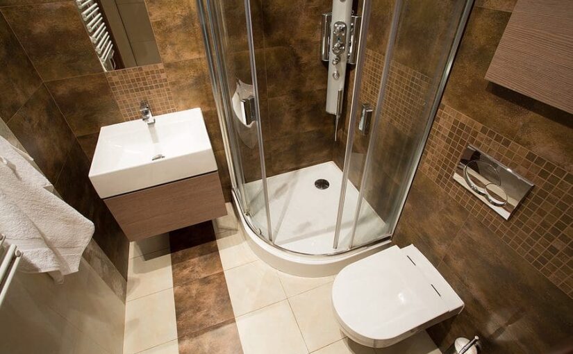 Modern small bathroom with brown tile walls, white curved shower enclosure, wall-mounted sink with wood vanity, and white toilet on cream and brown checkered floor