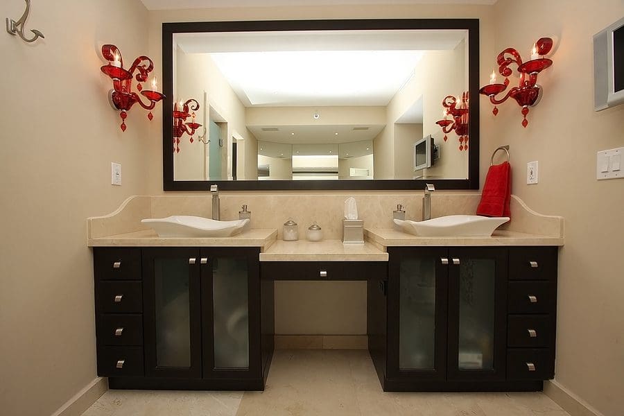 Luxury double vanity bathroom with black cabinetry, beige countertops, dual white vessel sinks, large framed mirror, and red decorative sconces