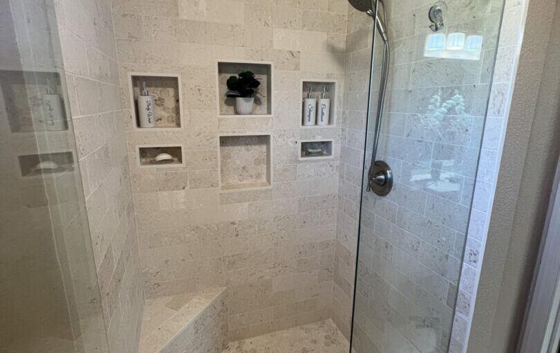 Modern luxury bathroom shower with travertine tile walls, built-in niches, rainfall showerhead, and frameless glass enclosure
