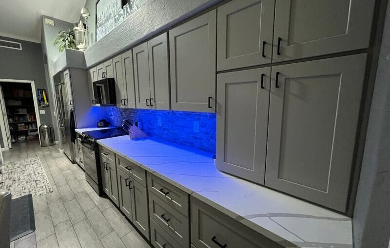 Modern kitchen with gray cabinetry, blue illuminated countertops, and stainless steel appliances under bright overhead lighting