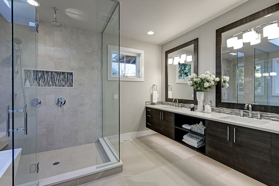 Spacious modern bathroom with gray tile walk-in shower, dark wood double vanity with white countertop, large mirrors, white flowers, and skylights