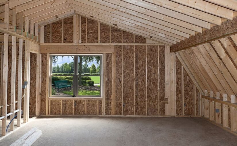 Interior of new room addition under construction with wooden frame, insulation, pitched ceiling beams, and large window overlooking green garden and lawn
