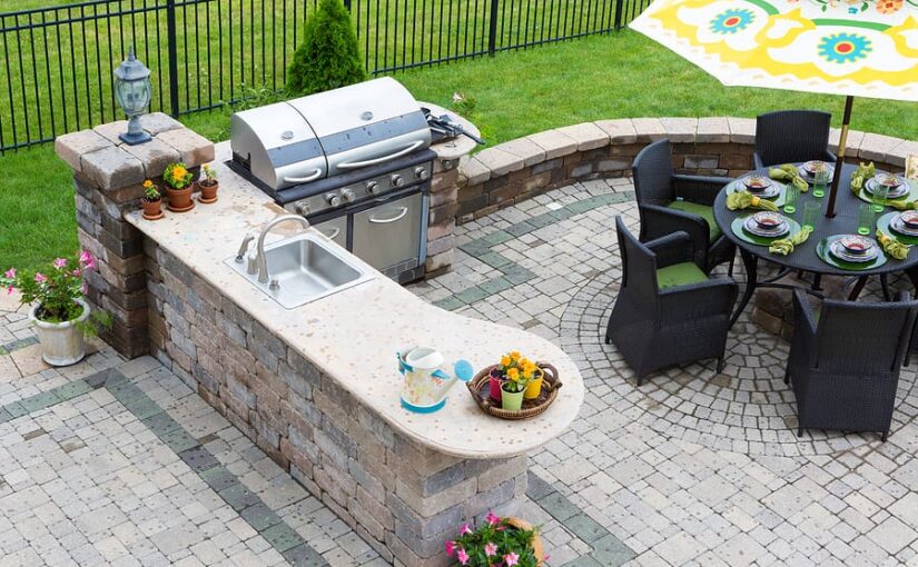 Outdoor kitchen and dining patio featuring a built-in gas grill, sink counter, and round dining table with place settings on brick pavers