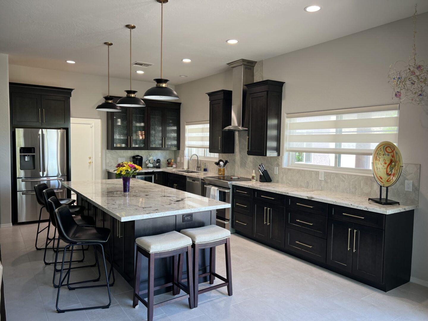 Modern kitchen with black cabinetry, marble countertops, stainless steel appliances, pendant lights over island with seating, and light gray walls