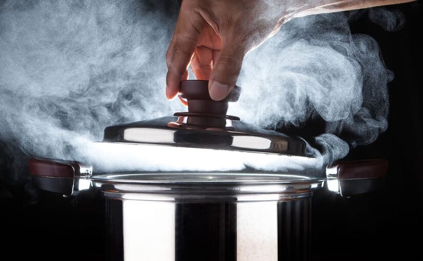 Hand of chef opening a steaming pressure cooker lid with dramatic studio lighting against a black background