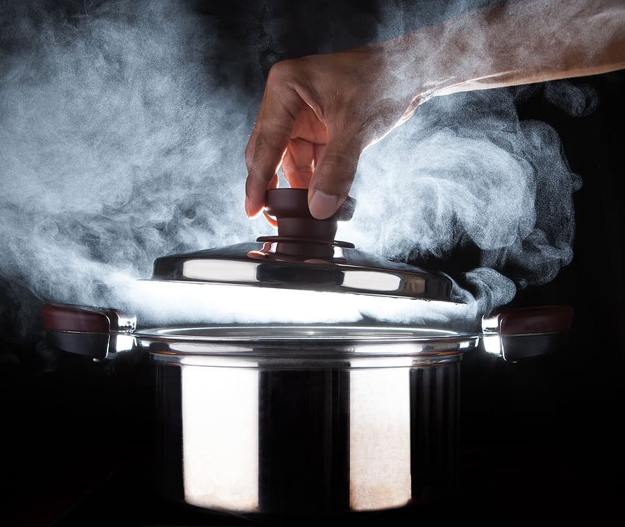 Hand of chef opening a steaming pressure cooker lid with dramatic studio lighting against a black background