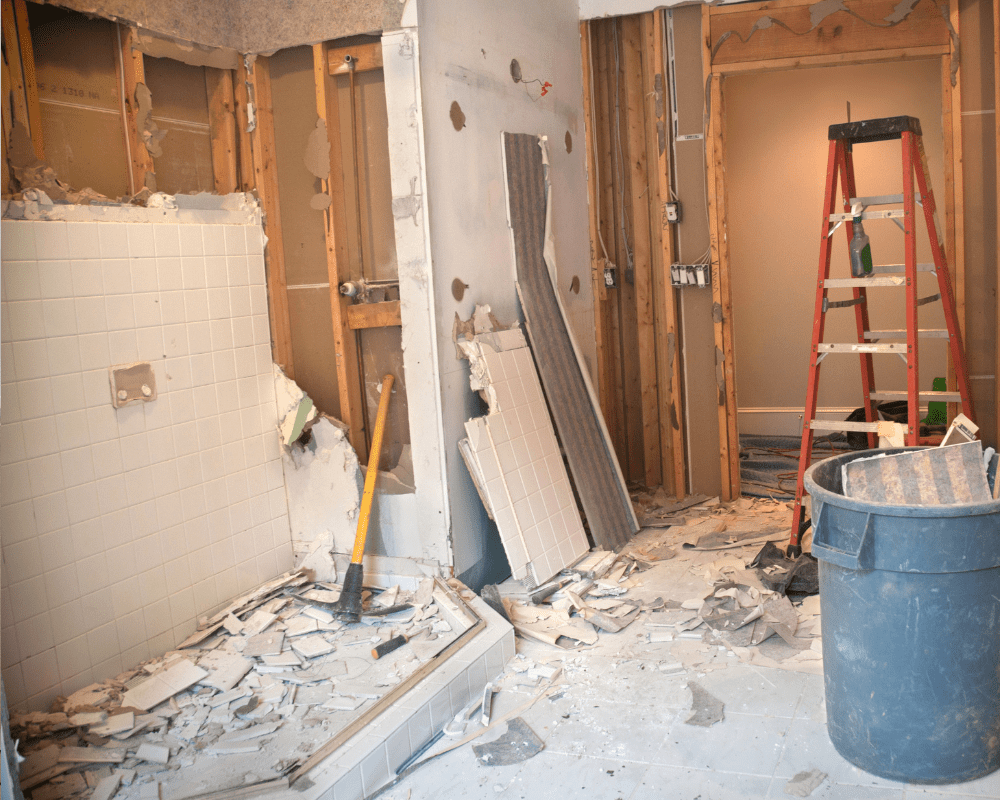 Bathroom renovation in progress with exposed framing, removed fixtures, scattered debris, a red ladder, and blue utility bucket