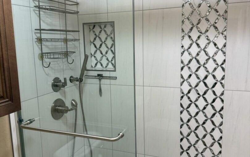 Modern bathroom shower enclosure with glass doors, rainfall showerhead, dual controls, metal shelving, and decorative black and white diamond tile accent strip on white subway tile walls