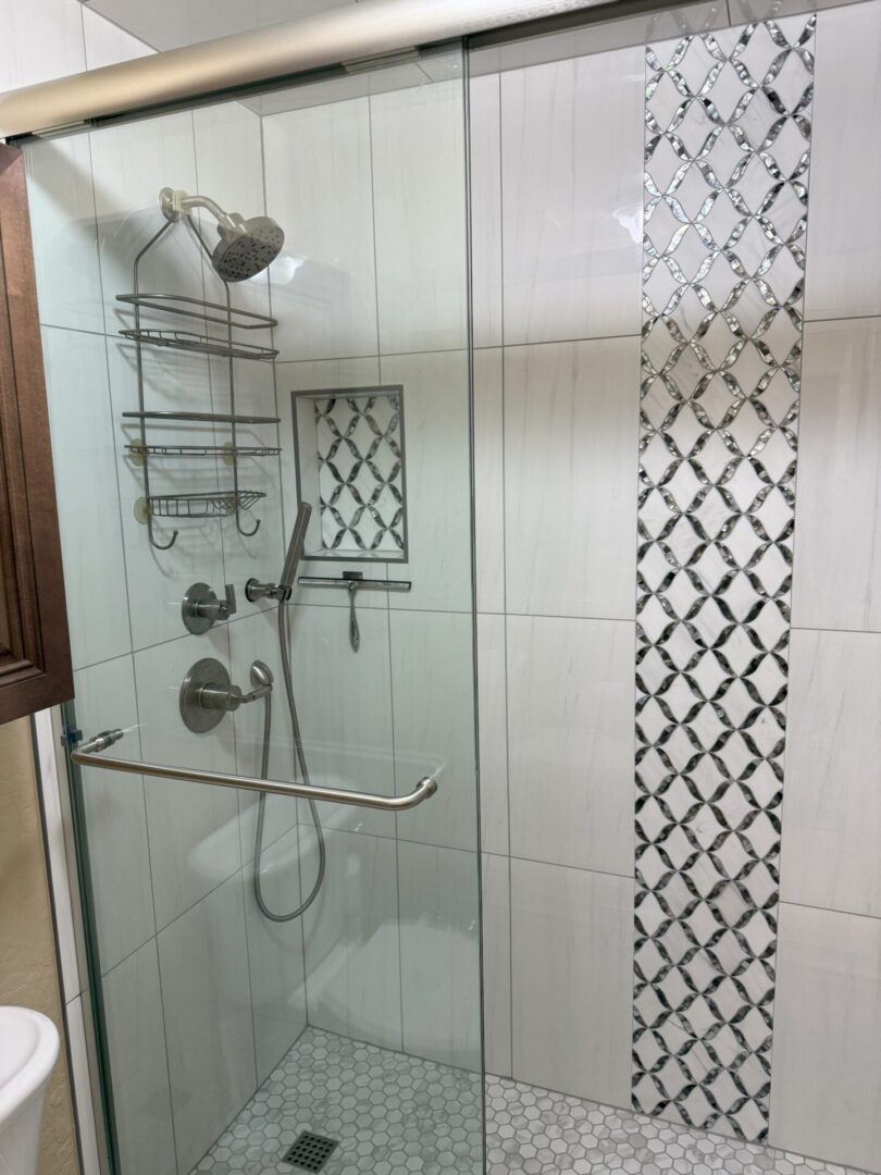 Modern bathroom shower enclosure with glass doors, rainfall showerhead, dual controls, metal shelving, and decorative black and white diamond tile accent strip on white subway tile walls