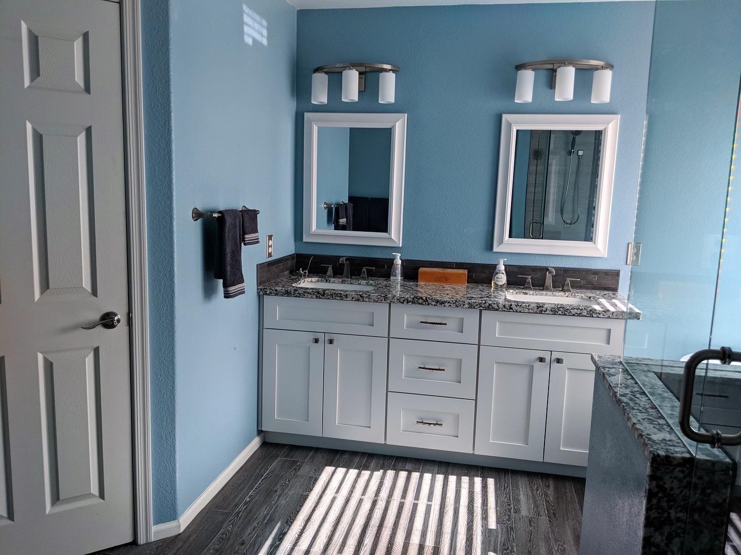 Modern bathroom with light blue walls, white framed mirrors with sconces above double granite vanity, white cabinetry, and dark wood-look flooring