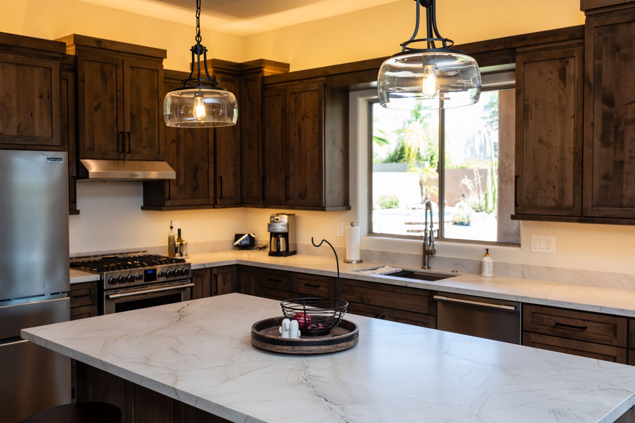 Modern kitchen with dark wood cabinetry, marble countertops, stainless steel appliances, and pendant lighting overlooking backyard patio