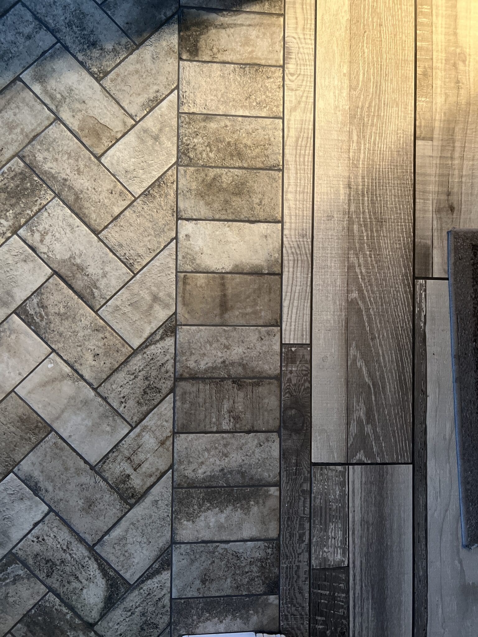 Four flooring samples displayed side-by-side showing herringbone tile, rectangular tile, dark wood plank, and light wood plank patterns