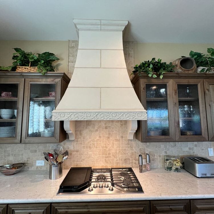 Elegant kitchen with cream tile range hood, gas cooktop, marble countertops, wood cabinetry with glass doors, and decorative plants on upper shelves