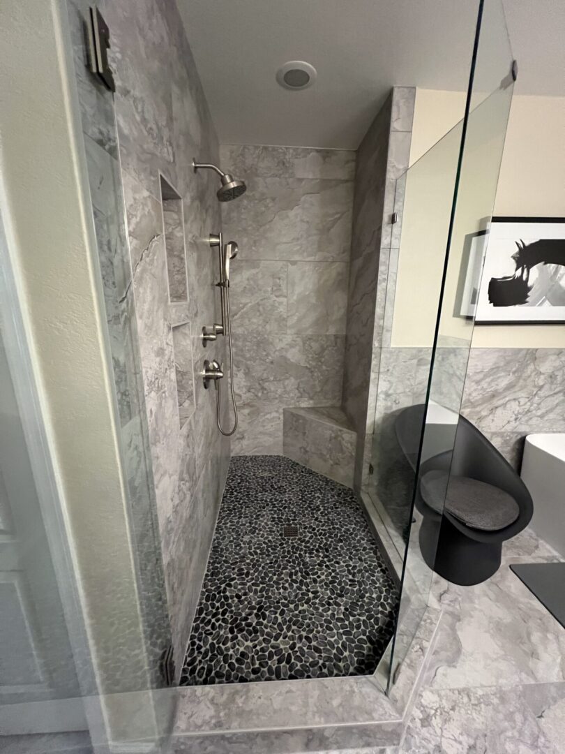 Modern bathroom shower with gray marble tile walls, black pebble stone floor, glass enclosure, and brushed nickel fixtures