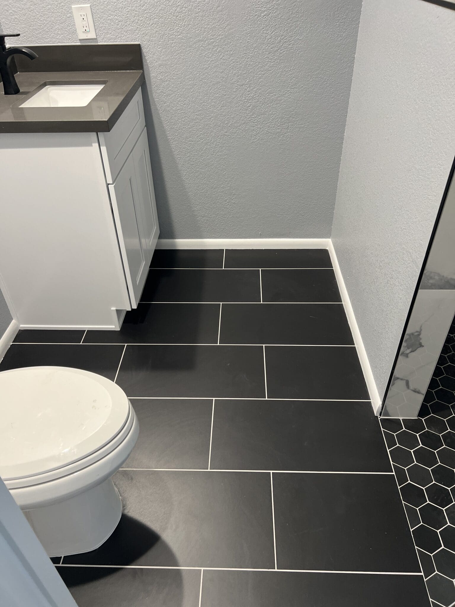 Modern bathroom with black subway tile flooring, white toilet, gray vanity cabinet, light gray walls, and black hexagonal tile accent in corner