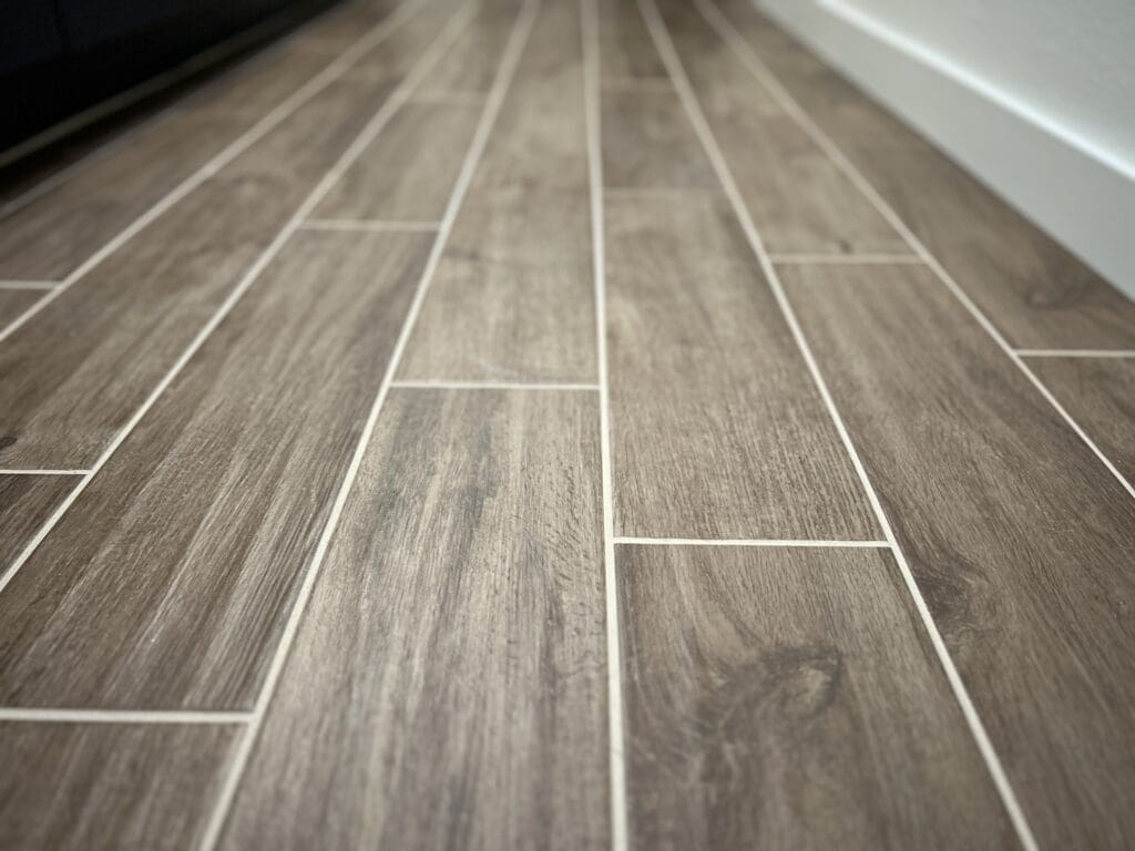 Close-up of gray wood-grain tile flooring with white grout lines in a herringbone pattern, showing realistic wood texture and realistic shadows