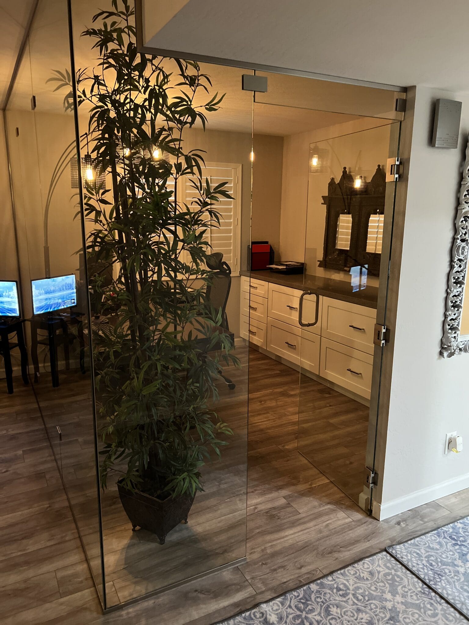 Modern spa bathroom with frameless glass shower enclosure, light wood vanity with dark countertop, bamboo plant, warm lighting, and wooden flooring