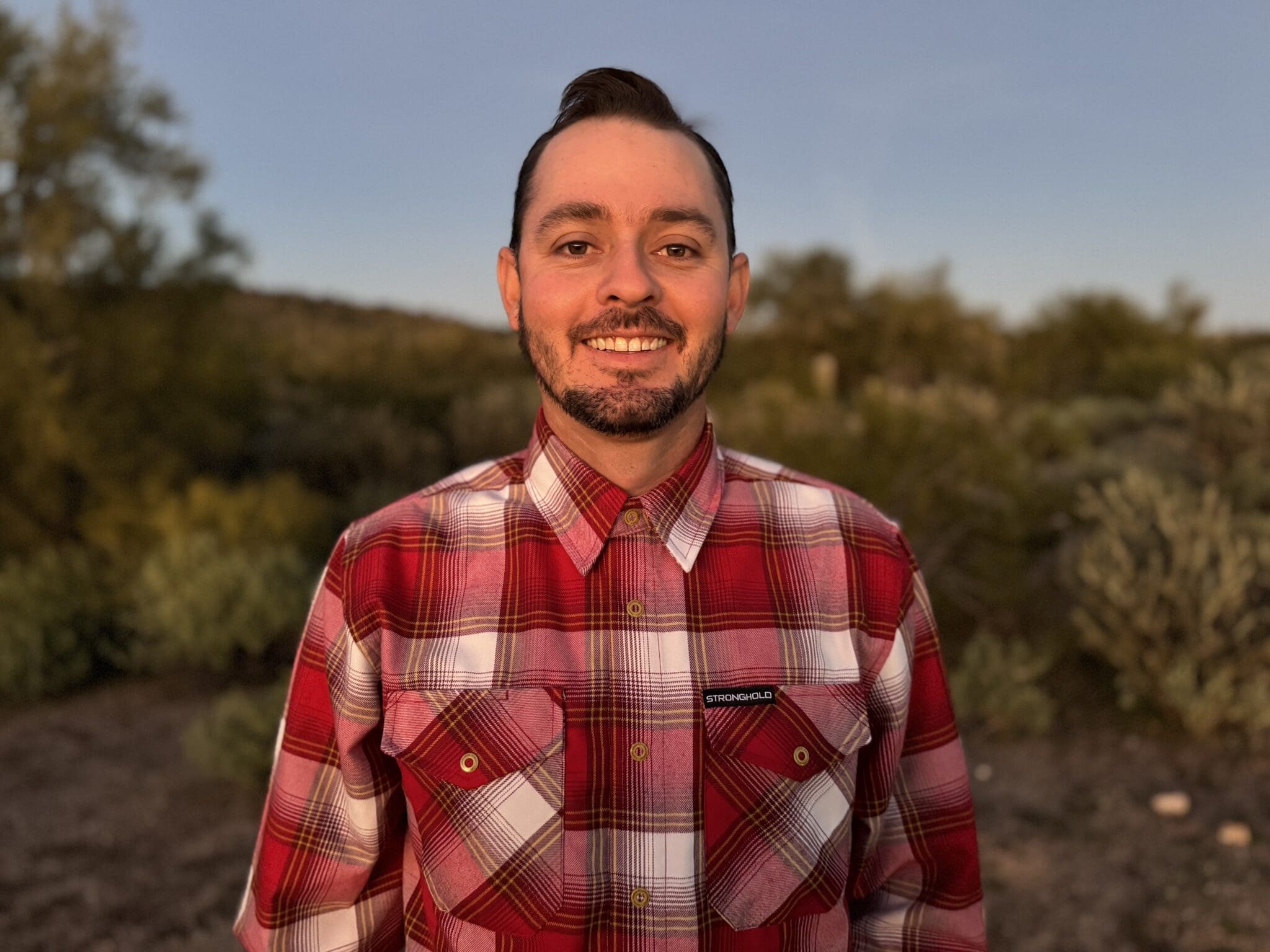 Man wearing red plaid shirt smiling at camera with desert brush landscape behind him
