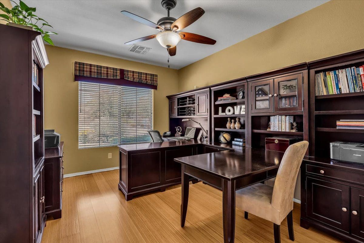 Spacious home office with dark wood desks, built-in bookshelves, cream walls, hardwood floors, and ceiling fan with natural light from shuttered window