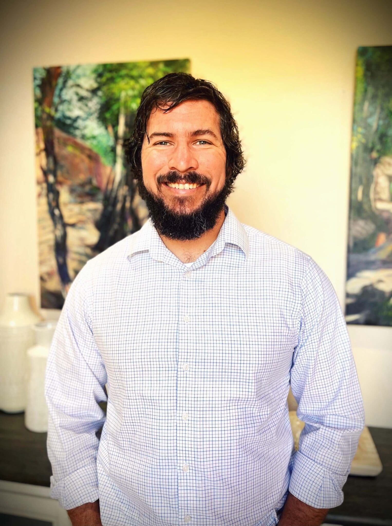 Justen Vickery, a bearded man with dark hair, smiling at the camera while wearing a light blue checkered button-up shirt in front of landscape artwork