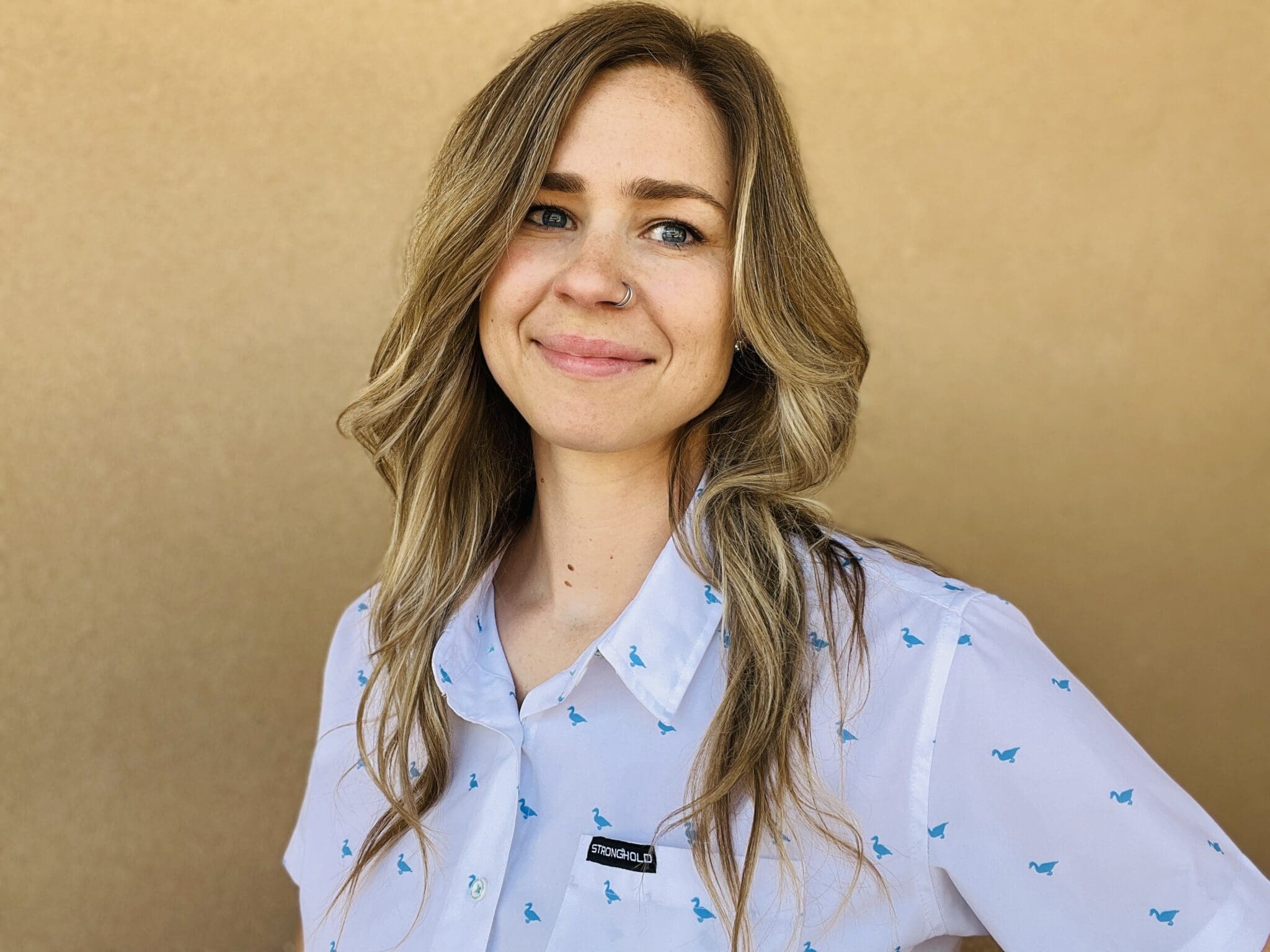 Woman with long brown wavy hair wearing a light blue collared shirt with blue bird pattern and small black logo badge, smiling against a tan background