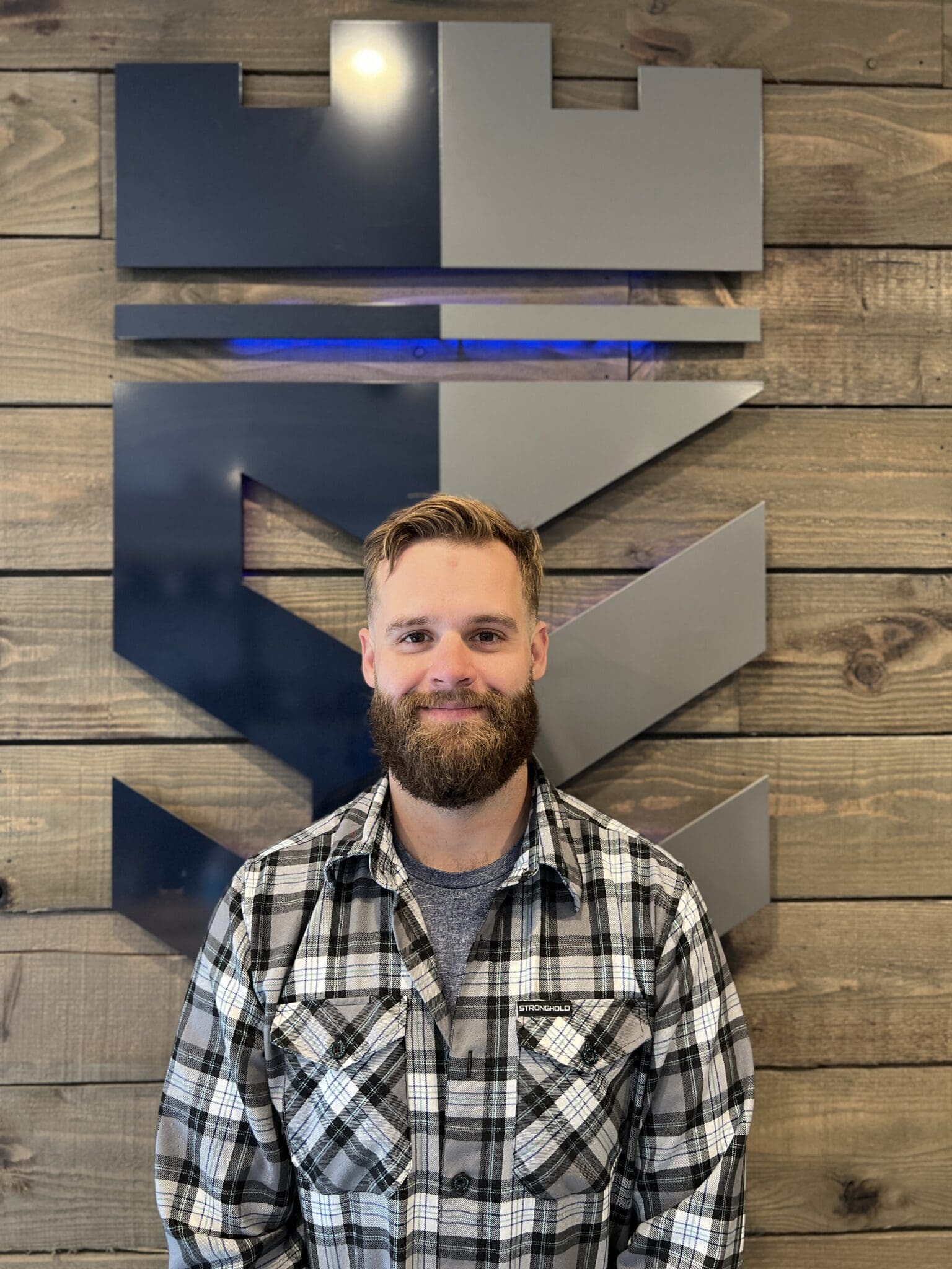 Man with beard wearing black and white plaid shirt standing in front of wooden wall with geometric blue and gray abstract art installation