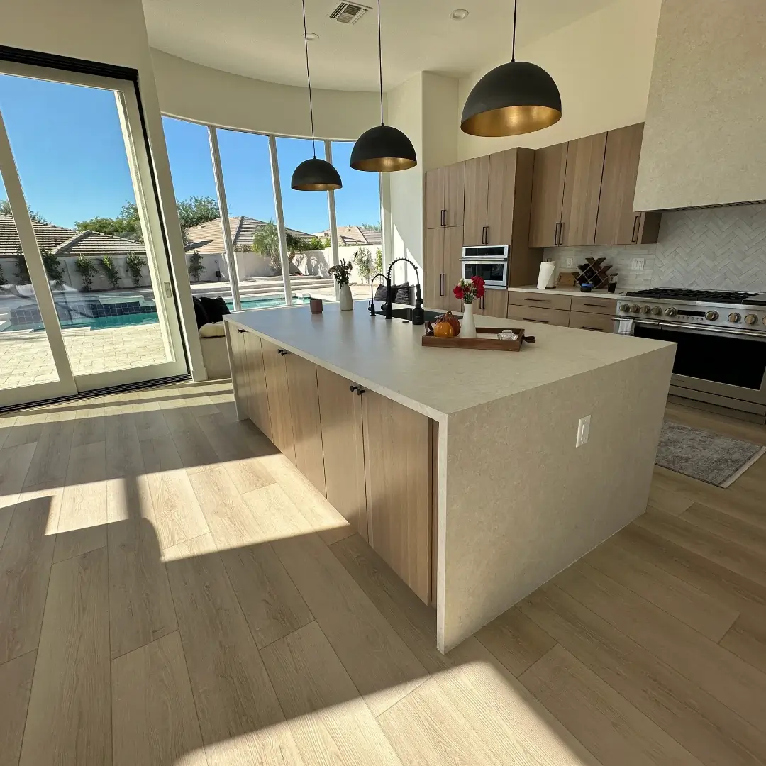 Modern kitchen with light wood cabinetry, white quartz island countertop, and black pendant lights overlooking a pool and patio through floor-to-ceiling windows