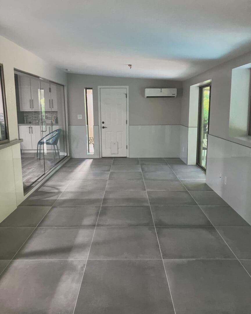 Spacious modern kitchen and dining area with gray tile flooring, white cabinetry, stainless steel appliances, and natural light from glass doors and windows